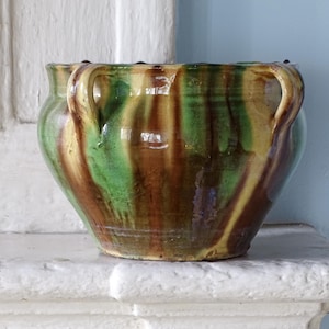 May include: A ceramic pot featuring a glossy glaze with vertical streaks of green, brown, and yellow. The vessel has a rounded body, a wide opening with a slightly scalloped rim, and two small, curved handles attached to the sides of the upper rim.