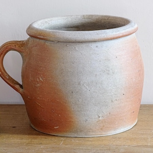 Op de afbeelding: Een rustieke keramische pot in antieke stijl met een handvat. De pot heeft een brede opening en een afgeronde body. De buitenkant heeft een tweekleurig kleurenschema, met een lichtgrijze bovenste helft en een roodbruine onderste helft. De pot staat op een houten oppervlak.