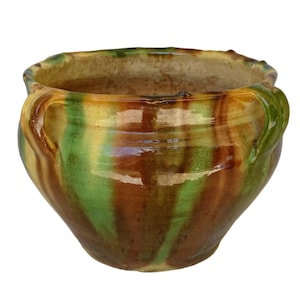 Op de afbeelding: Een handgemaakte keramische pot met een brede opening en twee handvatten. De pot heeft een mix van kleuren, waaronder groen, bruin en geel, met een glanzende afwerking. Dit decoratieve stuk is geschikt voor binnen of buiten.