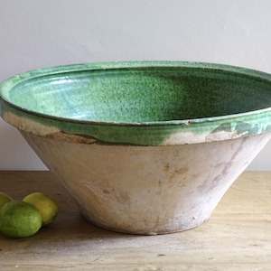 May include: A large, antique, off-white and green ceramic bowl with a wide rim. The bowl has a conical shape and a textured surface. Three green limes sit on a wooden surface in front of the bowl.