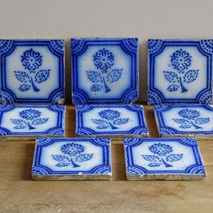 May include: Seven square ceramic tiles with a blue and white floral design. Each tile features a central flower and leaf motif, framed by a blue border with dotted accents. The tiles are arranged on a wooden surface.