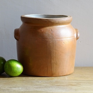 May include: A large, brown ceramic crock with two side handles and a slightly flared rim. The crock has a number "5" etched on the front. Three green limes sit in front of the crock on a wooden surface.
