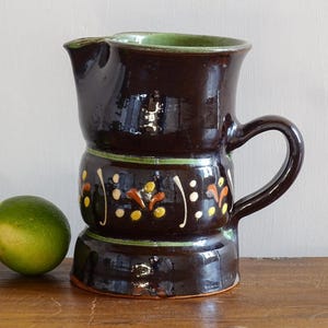 May include: A dark brown ceramic pitcher with a green rim and handle. The pitcher has a decorative band with colorful floral designs. Two limes are on the left side of the pitcher.