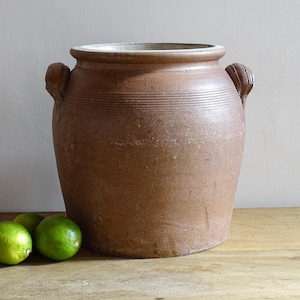 Puede incluir: Un jarrón grande de gres antiguo marrón con dos asas. El jarrón tiene una abertura ancha y una superficie texturizada con líneas horizontales. Tres limas verdes están frente al jarrón sobre una superficie de madera.