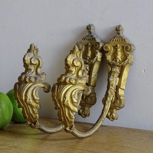 May include: A pair of gold-colored metal curtain tiebacks featuring ornate, scrolled leaf detailing and a vertical mounting plate. The hardware displays a weathered, textured finish with visible signs of age and wear across the metallic surface.