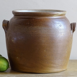 May include: A large, round, brown stoneware crock featuring a wide opening, a thick rim, and two small side handles. The exterior surface has a textured, earthy finish with subtle horizontal grooved lines encircling the upper body.