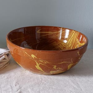 May include: A large, round ceramic mixing bowl with a glossy finish. The exterior and interior feature a marbled pattern of deep terracotta brown and bright yellow swirls, creating a fluid, organic design across the entire surface of the bowl.