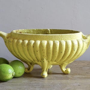Puede incluir: Un cuenco de hierro fundido amarillo con exterior acanalado, dos asas laterales y tres patas, con un acabado de pintura desgastado. Dos limas de color verde brillante están situadas a la izquierda del cuenco, que tiene una boca ancha y textura.