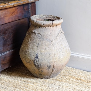 May include: An aged, beige earthenware pot with a textured surface and a wide body. The pot features decorative detailing around the neck and sides. It is placed on a woven jute rug next to a dark wooden cabinet. The pot's height is approximately 50 cm.