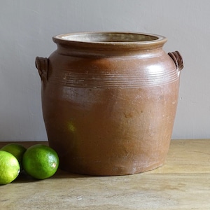 May include: A large, brown antique stoneware crock with two side handles. The crock has a wide opening and a slightly lighter-coloured rim. Three green limes sit on a wooden surface in front of the crock.