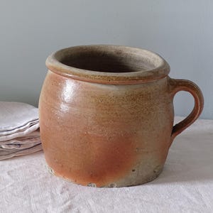 Może przedstawiać: Okrągły, beżowy garnek ceramiczny z pojedynczym uchwytem z boku i szerokim otworem. Zewnętrzna powierzchnia posiada nakrapianą, ziemistą glazurę z różnicami w odcieniach, od jasnego beżu po głębszy, wypalany pomarańczowy kolor z teksturą.