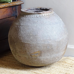 May include: A large, round, antique ceramic jar with a wide opening and a textured, weathered surface. The jar is a muted grey-brown colour and sits on a woven rug. The rim has a decorative scalloped edge.