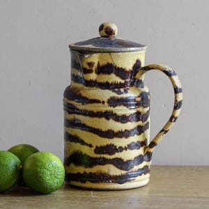 Può includere: Una brocca in ceramica con base gialla e strisce orizzontali marrone scuro screziate. La brocca presenta un coperchio abbinato con una piccola maniglia a pomello, un manico laterale curvo e un beccuccio. Tre lime verde brillante a sinistra.