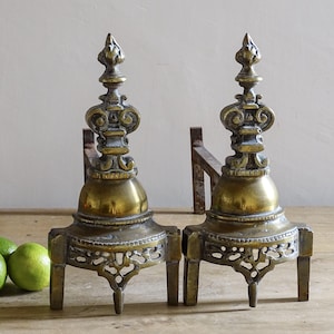 Peut inclure: Deux chenets anciens en laiton avec des détails ornés. Chaque chenet présente un fleuron décoratif, un corps bulbeux et une base ajourée. Les chenets sont exposés sur une surface en bois avec trois citrons verts.