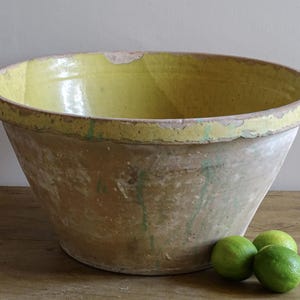May include: A large, round, earthenware mixing bowl with a wide, flared rim. The interior is glazed in a bright yellow, while the exterior features a weathered, unglazed terracotta finish with visible green paint drips and signs of wear.