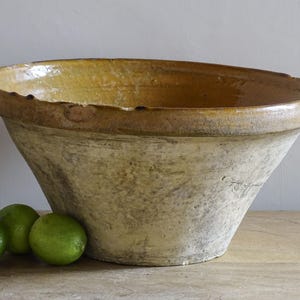 May include: A large, antique-style ceramic bowl with a wide rim and a light yellow interior. The exterior has a textured, aged appearance. Three green limes rest in front of the bowl, suggesting a kitchen or food-related theme.