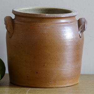 May include: A cylindrical brown stoneware crock featuring a wide mouth, a thick rim, and two small, vertically ribbed side handles. The exterior surface displays a mottled, earthy texture with subtle horizontal incised lines around the body.