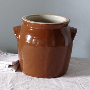 May include: A brown glazed ceramic crock with a wide circular opening, a thick rim, and two small side handles. The exterior features a smooth, glossy finish in a deep earthy brown tone, while the interior is a lighter, neutral off-white colour.