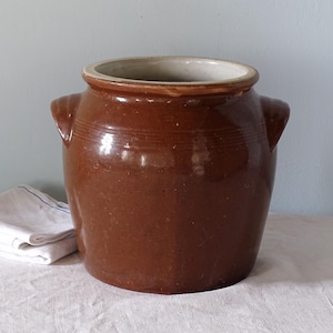 May include: A brown glazed ceramic crock with a wide circular opening, a thick rim, and two side handles. The exterior features a smooth, glossy finish with subtle horizontal ridges near the top, while the interior is a light, unglazed beige colour.