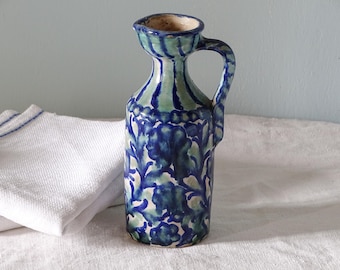 May include: A ceramic pitcher featuring a hand-painted floral pattern in shades of deep blue and light green. The vessel has a cylindrical body, a narrow neck with a flared rim, and a single curved handle extending from the neck to the shoulder.
