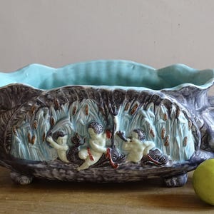 Può includere: Un grande vaso rettangolare in ceramica con interno azzurro e esterno grigio scuro. Il vaso presenta un disegno a rilievo di cherubini e canne. Il vaso poggia su quattro piedini.