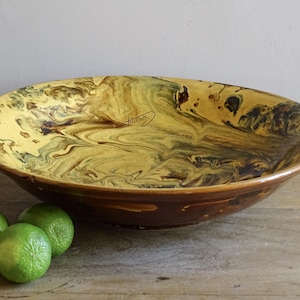 Peut inclure: Un grand bol en céramique peu profond avec un vernis marbré dans des tons de jaune, brun et vert foncé. La surface intérieure présente un motif fluide et tourbillonnant avec une signature manuscrite. Trois citrons verts sont posés à côté.