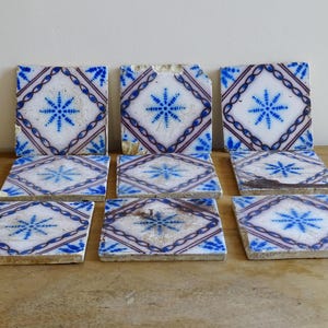 May include: Nine antique square tiles with a white background, blue starburst design, and diamond pattern. The tiles have a distressed appearance, with some chipped edges and wear. The tiles are arranged on a wooden surface.