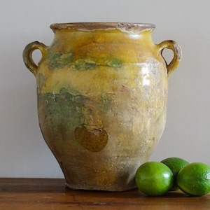 Può includere: Un grande vaso in ceramica usurata con due manici laterali, caratterizzato da una finitura smaltata gialla e verde maculata con texture superficiale visibile e segni del tempo. Tre lime verdi sono posizionate davanti al vaso.