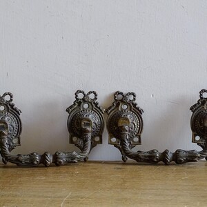 Peut inclure: Deux poignées de tiroir en laiton de style ancien assorties, dotées de volutes détaillées et de plaques décoratives avec des sommets en forme de nœud. Chaque poignée présente un design oscillant et texturé avec des motifs en relief sombres.