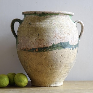 Könnte beinhalten: Eine große, gealterte Vase aus beige-grüner Keramik mit zwei Henkeln. Die Vase hat eine strukturierte Oberfläche mit einem grünen Band in der Mitte und am oberen Rand. Drei grüne Limetten liegen vor der Vase.