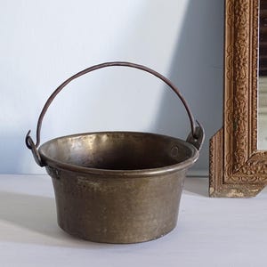 May include: A round, weathered brass bucket with a dark, oxidized patina and a textured, hammered surface. It features a thin, arched metal handle attached to two side loops, creating a simple, functional vessel with visible signs of age.