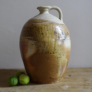 Peut inclure: Une grande cruche en céramique bombée avec une anse unique et un col étroit. Le récipient présente une glaçure tachetée dans des tons de beige, brun et crème, avec une finition terreuse texturée. Trois petits citrons verts ronds sont posés.