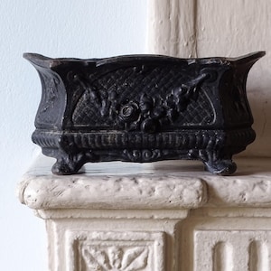 May include: A rectangular, dark brown cast iron planter featuring an ornate, raised floral relief pattern on the front and sides. The vessel has a textured, cross-hatched background, a scalloped top edge, and rests on four small, decorative feet.