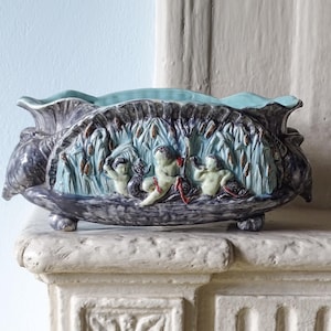May include: An oval ceramic planter with a dark, mottled glaze and a light blue interior. The exterior features a relief sculpture of three figures playing among tall reeds and cattails, supported by small, rounded feet at the base.