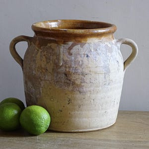Może przedstawiać: Duży, ceramiczny garnek w stylu antycznym z dwoma uchwytami, kremowym korpusem i brązową krawędzią. Garnek ma wygląd postarzanego, z widocznymi niedoskonałościami. Trzy zielone limonki leżą przed garnkiem na drewnianej powierzchni.