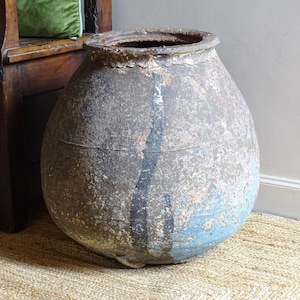 May include: Large, weathered, grey and blue ceramic pot with a wide mouth. The pot has a rounded body and a textured surface. A dark vertical stripe runs down the front. The pot is on a woven rug.