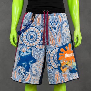 May include: Knee-length shorts featuring a white base with a blue and orange paisley pattern. The shorts include a dark blue star patch, a Nickelodeon logo with cartoon characters, a black waistband with magenta drawstrings, and a blue chain.