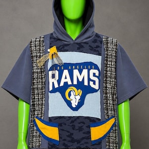 May include: A short-sleeved hooded sweatshirt in slate blue featuring a central Los Angeles Rams logo patch. The garment includes side panels of black and white tweed, two yellow and blue pockets, and a dark blue camouflage pattern.