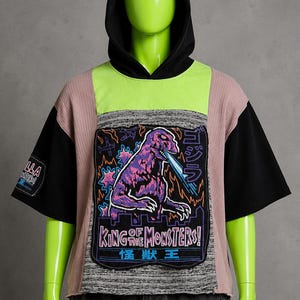 May include: A short-sleeve hooded shirt featuring a black hood, neon green chest panel, and mauve textured fabric. The center graphic depicts a purple Godzilla with blue breath, the text "KING OF THE MONSTERS!", and Japanese characters.