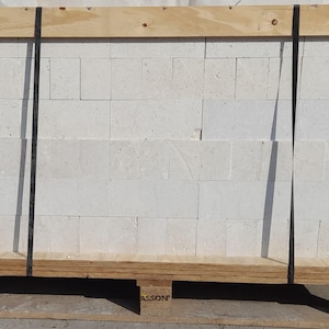 May include: A pallet of white concrete blocks, stacked and secured with black straps. The blocks are arranged in a brick pattern. The pallet is made of wood and has a label that reads "Silresim Calcia Cemento".