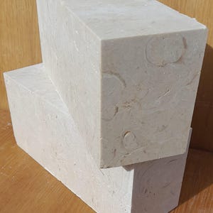 May include: Two rectangular blocks of light beige travertine stone. The blocks are stacked on top of each other. The stone has a natural, textured surface with a subtle pattern of swirls and veins.