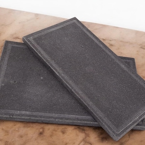 Peut inclure: Deux plateaux de service rectangulaires en ardoise noire avec une bordure légèrement surélevée. Les plateaux sont empilés l'un sur l'autre.