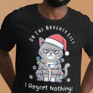 May include: Black t-shirt featuring a grumpy gray cat wearing a Santa hat and wrapped in Christmas lights. The text "ON THE NAUGHTY LIST" arches above the cat, with "I Regret Nothing!" below.