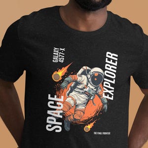 May include: Dark grey t-shirt with a graphic of an astronaut in a spacesuit, the words "SPACE EXPLORER" and "GALAXY 4577-X". The astronaut is surrounded by orange flames and a meteor. The text "THE FINAL FRONTIER" is also present.