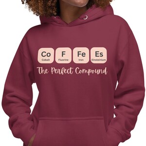 Könnte beinhalten: Ein weinroter Kapuzenpullover mit einer Vordertasche und einer Kapuze mit Kordelzug. Auf der Vorderseite des Kapuzenpullovers steht der Text "Co F Fe Es" im Stil eines Periodensystems, darunter die Worte "The Perfect Compound".