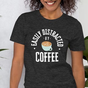 Può includere: Una t-shirt grigio scuro melange con una grafica bianca che dice "Easily Distracted by Coffee" con un'immagine di cartone animato di una tazza di caffè.