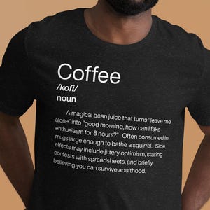 Peut inclure: Un t-shirt gris foncé avec le mot "Coffee" et sa prononciation phonétique en blanc. En dessous, une définition humoristique du café est imprimée en blanc. Le t-shirt est porté par une personne souriant à la caméra.