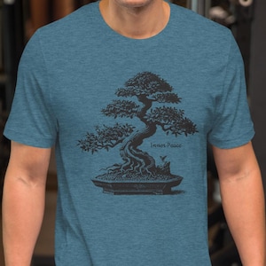 May include: Teal short-sleeve t-shirt featuring a black illustration of a bonsai tree in a pot. The tree has a detailed, stylized design with the words "Inner Peace" below it. The shirt is made of a soft material.
