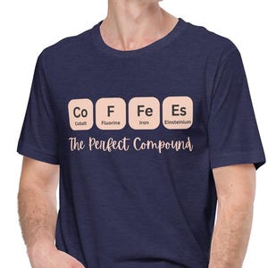 Può includere: T-shirt blu navy con un design della tavola periodica che scrive "COFFEE" usando gli elementi Cobalto, Fluoro, Ferro ed Einstenio. Il testo "The Perfect Compound" è sotto.