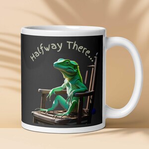 Könnte beinhalten: Weiße Keramiktasse mit einer grünen Eidechse in einem hölzernen Schaukelstuhl. Die Eidechse sitzt mit verschränkten Armen und Beinen. Die Tasse trägt den Text "Halfway There..." in weißer Schrift auf schwarzem Hintergrund.